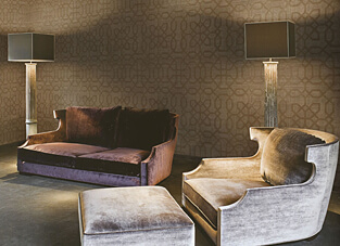 Ein elegant inszenierter Wohnbereich, dessen Wände mit einer edlen Velours-Tapete in einem dunklen Taupeton versehen sind. Die Tapete zeigt ein wiederkehrendes, geometrisches Muster in einem leichten Schimmer. Im Vordergrund stehen ein kleineres, dunkelviolettes Samtsofa und ein hellgrauer Samtsessel mit Ottoman, flankiert von zwei hohen Stehlampen.