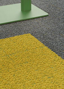 Nahaufnahme eines Bodens, der mit hoch strapazierfähigen "Iconic" Teppichfliesen von "esso" ausgelegt ist. Der Hauptbodenbelag ist eine graue Fliese. Eine weitere Fliese in leuchtendem Gelb dient zur Zonierung von Flächen. Beide Teppichfliesen bestehen aus 100% nachhaltigem Econyl und liegen auf einem Ecobase-Träger. Im Hintergrund ist die Basis eines modernen, hellgrünen Möbels zu sehen.