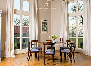 Gerne, hier ist der Alt-Text mit dem Fokus auf die Wandfarbe Pemberley von Anna von Mangoldt: Alt-Text: Ein heller Essbereich in einem Altbau mit großen Fenstertüren. Die Wände sind in der hellen, warmen Farbe "Pemberley" von ANNA VON MANGOLDT FARBEN gestrichen. In der Raummitte stehen ein runder Holztisch und klassische Stühle mit blauen Polstern.
