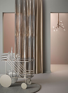 Eine moderne Interieurszene in hellen Grau- und Rosatönen. Rechts neben einem transparenten, metallisch glänzenden Vorhang hängt ein klassischer, glänzender Dekorationsstoff von NYA Nordiska in einem satten Gold-/Beigeton. Im Vordergrund steht ein eleganter, silberner Barwagen mit Glaswaren auf einem runden, grauen Teppich.