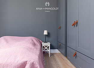 Ein Schlafzimmer, dessen Wände in einem matten, dunklen Graublau (Anthrazit oder Schiefergrau) gestrichen sind. Das graue Farbkonzept wird durch einen hellen Holzboden und einen großen Einbauschrank in einem passenden, aber etwas helleren Grauton ergänzt. Ein Bett mit einer rosa-pinken gesteppten Tagesdecke und braune Ledergriffe an den Schranktüren setzen warme Kontraste. Das Logo von ANNA VON MANGOLDT FARBEN ist in Weiß oben an der Wand platziert.