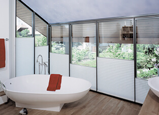 Ein modernes Badezimmer mit einer freistehenden, weißen Oval-Badewanne vor einer großflächigen, schrägen Fensterfront. Die Fenster sind mit einer weißen Waben-Plissee-Anlage versehen, die von unten nach oben verstellbar ist, um sowohl Sichtschutz als auch gute Tageslichtnutzung zu gewährleisten. Ein rotes Handtuch liegt auf dem Badewannenrand.