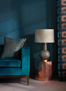 Nahaufnahme eines Interieurs mit einer Wand in einem tiefen Blau-Türkis. Die Armlehne und das Kissen eines Sofas sind mit einem klassischen Velours-Stoff von Osborne & Little in Türkis bezogen. Das Kissen zeigt ein orange-blaues Zickzack-Muster. Neben dem Sofa steht eine Tischleuchte auf einem glänzenden Kupferzylinder.