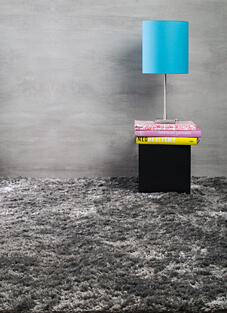 Raum mit grauer Betonwand und luxuriösem Langflor-Teppich in dunklem meliertem Grau. An der Wand ein kleiner schwarzer Beistelltisch mit zwei Büchern und einer türkisfarbenen Lampe darauf.