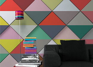 Designbeispiel modernes Wandpanel. Wand bestehend aus verschieden farbigen Dreiecken. Davor ein schwarzer Sessel, Bücherstapel und eine geradlinige bunt gestreifte Stehlampe.