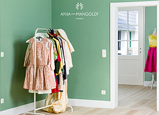 Ein helles Zimmer, gestrichen in einem matten Salbei- oder Minzgrün mit weißen Sockel- und Türleisten. Rechts steht ein weißer Kleiderständer mit verschiedenen Kinderkleidern und Kostümen. Über dem Kleiderständer ist das Logo von ANNA VON MANGOLDT FARBEN platziert. Durch die offene, weiße Tür ist ein weiterer Raum mit einer Schaufensterpuppe in einem leuchtend pinken Kleid sichtbar.