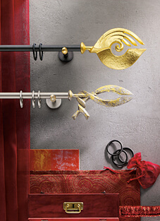 Designbeispiel von zwei Gardinenstangen mit Ringen daran, montiert auf einer hellgrauen Wand mit jeweils farblich passenden Haltern. Oben schwarze Stange mit kunstvollem goldenen Endstück, unten silberne Stange mit Silber/goldenem kunstvollem Endstück.