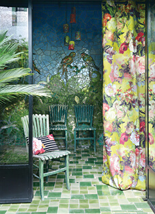 Eine kleine, helle Veranda oder ein Wintergarten mit einem Boden aus grünen Kacheln und zwei grünen Holzstühlen. Rechts hängt ein Dekorationsstoff von Lelièvre im romantischen Landhausstil, der ein großflächiges, florales Muster aus Blumen in Rot, Pink und Grün auf einem leuchtend gelb-grünen Hintergrund zeigt. Die Rückwand ist mit einem Mosaik aus türkisen und blauen Steinen mit Vogelmotiven verziert.