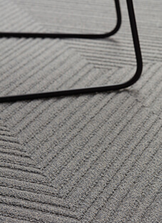 Nahaufnahme einer grauen, texturierten Teppichoberfläche, geometrische Teppichplanke mit einem erhabenen, gewinkelten Streifenmuster zu sehen. Auf dem Boden stehend die schwarzen, gebogenen Beine eines modernen Stuhls oder Tisches.