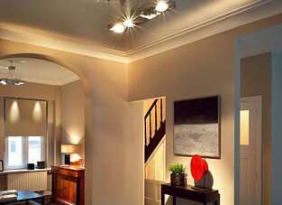 Ein Wohnbereich mit beigefarbenen Wänden und indirekter Beleuchtung, wobei klassische, weiße Deckenleisten die Übergänge von der Wand zur Decke schmücken.