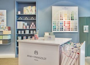 Ausschnitt des Shops von Anna von Mangold Farben. Hellblaue Wand im Hintergrund mit einem Regal befüllt mit Farben und einer Farbtafel daneben. Im Vordergrund eine weiße Kommode auf Rollen.