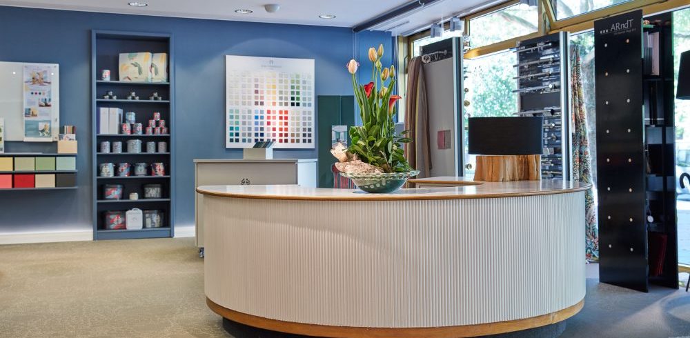 Heller Verkaufstresen von Anna von Mangold Farben mit großem Tulpenarrangement darauf. Im Hintergrund eine blaue Wand mit einem Einbauregal und einer großen Farbtafel. Rechts hinter dem Verkaufstresen eine raumhohe Fensterfront mit ausgestellten Gardinenstangen.