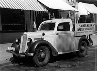 Schwarz-Weiß-Foto eines historischen, kleinen Lieferwagens (Pickup-Truck) aus dem Jahr 1950, den Zimmermann für 400 Mark erwarb. Auf der Seitenwand der Ladefläche steht "ZIMMERMANN TAPETEN-SPEZIALHAUS" in dunkler Schrift. Das Fahrzeug ist vor einer Geschäftsfassade mit gestreiften Markisen geparkt.