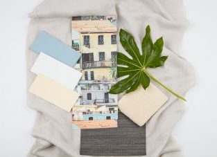 Ein Moodboard mit einer Sammlung von Stoffmustern, Farbkarten und einem Foto auf einem hellen Hintergrund. Es liegt ein Foto von einer mediterranen Häuserfassade mit Balkonen und Fensterläden in Pastelltönen. Daneben befinden sich Farbkarten in Hellblau, Weiß und Beige, ein kleines Stück beiger Stoff, ein dunkler Stoff oder Teppich sowie ein grünes, palmenartiges Blatt. Das Ganze ist auf einem hellgrauen Leinenstoff arrangiert.