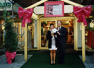 Historisches Foto aus dem Jahr 1999 anlässlich des 50. Firmenjubiläums von Zimmermann Interieur. Die Inhaber stehen vor dem dekorierten Ladeneingang, der von einem großen, rosa Schild und zwei großen, pinken Schleifen flankiert wird. Die Frau trägt ein weiß-schwarzes Kleid und der Mann einen dunklen Anzug. Ein grüner Teppich führt zur Tür.