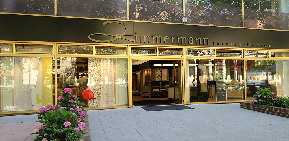 Außenansicht von Zimmermann InteriorDesign Showroom. Gläserne Fassade mit goldenen Einfassungen. Über dem Eingang schwarz abgesetzter Steifen mit der goldenen Aufschrift: "Zimmermann InteriorDesign". In den Schaufenstern verschiedene ausgestellte Vorhangstoffe. Recht und Links neben dem Eingang blühende Hortensien.