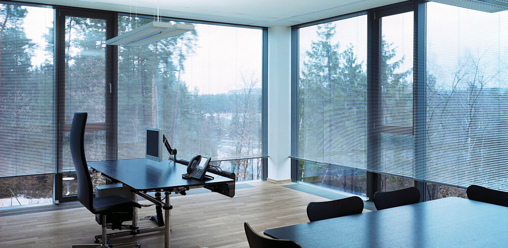 Designbeispiel für Sonnenschutz. Modern eingerichteter Büroraum mit Fensterfront über eine Ecke und weißen Plissees vor den Fenstern.