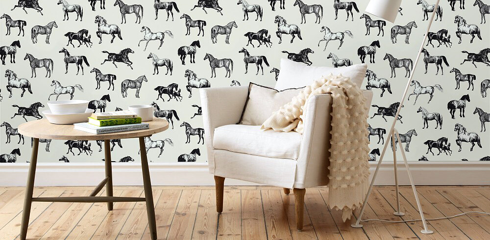 Designbeispiel einer Tapete. Wand mit weißem Untergrund und schwarzen Abbildungen von Pferden. davor auf hellem Holzboden ein kleiner hölzerner Beistellstich mit Büchern und Geschirr darauf. Rechts weißer Sessel mit Kissen und Decke darauf. Rechts eine moderne weiße Stehlampe.