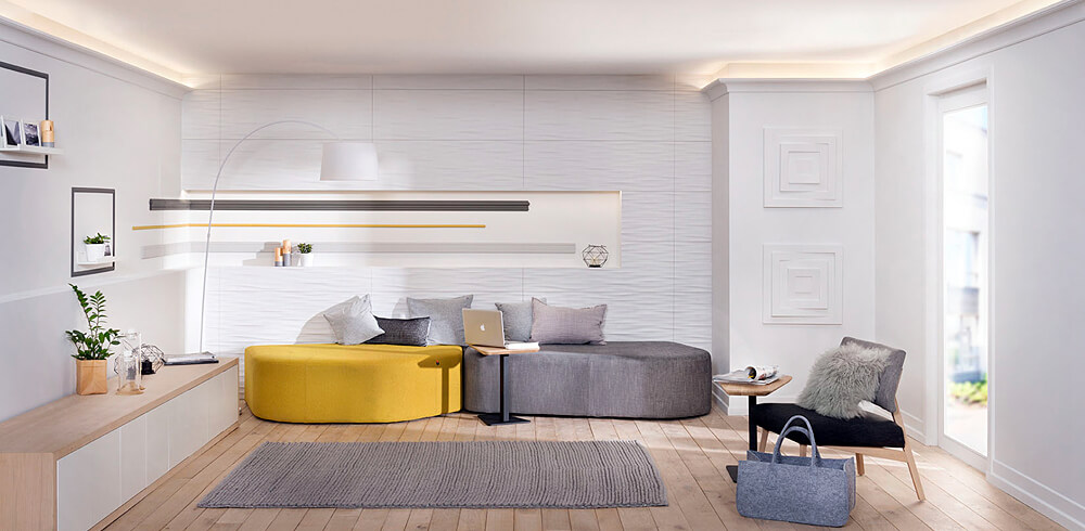 Hell gestalteter Wohnraum mit weißen Wänden und weißer Decke. rechts und links an den Wänden indirekt beleuchtete Zierleisten. Mittig ein grauer Teppich auf hellem Holzboden. Im Hintergrund eine moderne gelb Graue Couch, mit Kissen in verschiedenen Grautönen. Links ein Sideboard und recht ein gemütlicher grau schwarzer Sessel.