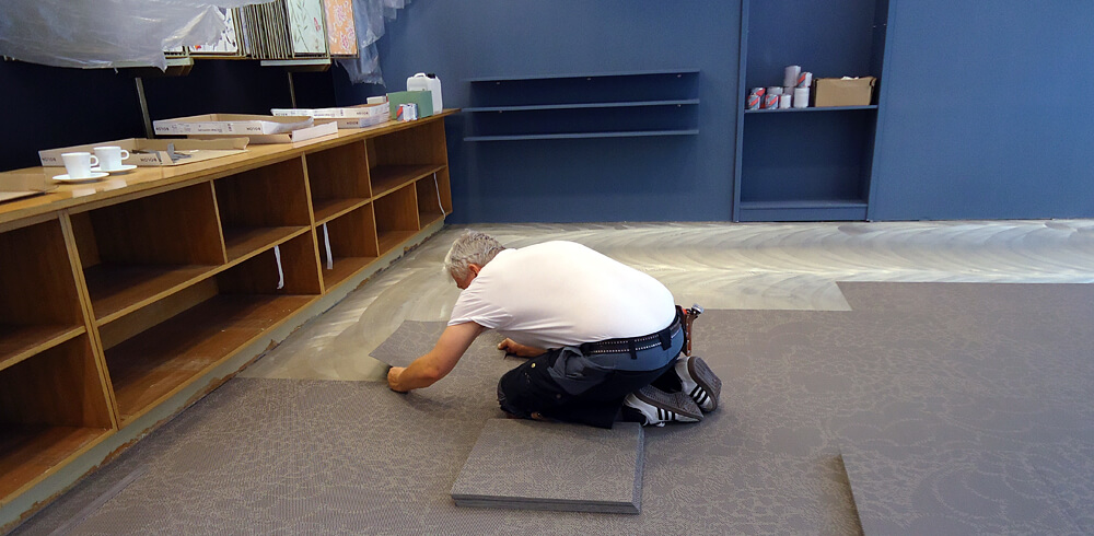 Bodenleger in blauer Arbeitshose, weißen Sneaker und weißem T-Shirt verlegt taupe Teppichfliesen. Links ein Sideboard und im Hintergrund eine blaue Wand mit Einbauregal.