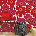 Designbeispiel von finnisch MARIMEKKO Tapete. Wand mit hellem Untergrund und großen roten und pinken Blumen darauf. Davor auf einem Holzpodest eine bunte Bank mit einer roten Tasche darauf. Rechts daneben eine weiß schwarz gestreifte große Tasche.