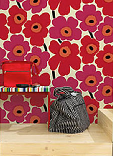 Designbeispiel von finnisch MARIMEKKO Tapete. Wand mit hellem Untergrund und großen roten und pinken Blumen darauf. Davor auf einem Holzpodest eine bunte Bank mit einer roten Tasche darauf. Rechts daneben eine weiß schwarz gestreifte große Tasche.
