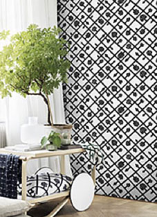 Tapetendesign-Beispiel von Marimekko Vol.5-03. Weiße Tapete mit schwarzem grafischen Muster darauf. Davor ein Servierwagen mit einer Pflanze darauf. Kissen darunter und Decke auf Schiebgriff drapiert.