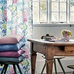 Designbeispiel von Osborne und Little Italienflair. Raumausschnitt im skandinavischen Stil. Im Hintergrund eine Fensterfront mit weißer Holzverkleidung. Rechts ein floraler Vorhang in blau Pink Grüntönen gehalten. Vor dem Vorhang ein Holzstuhl mit 4 gestapelten Kissen in Grün, Pink, Blau und Orange. Rechts im Vordergrund ein Holzschreibtisch mit einem türkisen Stuhl davor.