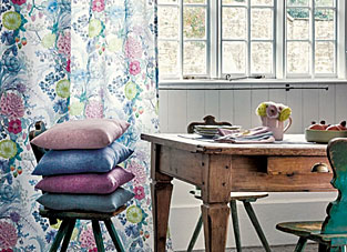 Designbeispiel von Osborne und Little Italienflair. Raumausschnitt im skandinavischen Stil. Im Hintergrund eine Fensterfront mit weißer Holzverkleidung. Rechts ein floraler Vorhang in blau Pink Grüntönen gehalten. Vor dem Vorhang ein Holzstuhl mit 4 gestapelten Kissen in Grün, Pink, Blau und Orange. Rechts im Vordergrund ein Holzschreibtisch mit einem türkisen Stuhl davor.