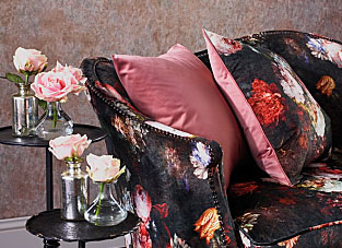 Designbeispiel der Firma Osborne and Little. Links eine Blumentreppe, dekoriert mit Rosen in kleinen gläsernen Vasen. Rechts ein dunkles Sofa mit floralem Muster. Auf dem Sofa zwei Kissen in stilvollem Rosa und auf der andern Seite im gleichen Muster wie das Sofa. Im Hintergrund eine Grau Bronze marmorierte Tapete.