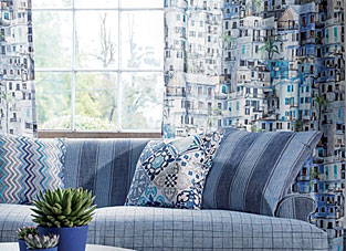 Designbeispiel von Osborne Little Italienflair. Fensterfront mit zwei Vorhängen in Blautönen mit Häusern darauf. Davor eine blaue Couch mit blauen Kissen. Im Vordergrund sind Sukkulenten.