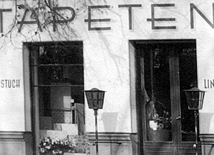 Schwarz-Weiß-Foto der Geschäftsfront von Zimmermann aus dem Jahr 1949 in Schmargendorf. Über den Fenstern ist in dunklen, serifenlosen Großbuchstaben der Schriftzug "TAPETEN" zu sehen. Vor dem Geschäft stehen zwei historische Straßenlaternen und ein kleines Blumenarrangement im Eingangsbereich.