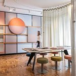 Moderner Showroom mit Vescom Akustikstoffen: Eine Rückwand zeigt farbige (Rosa, Hellblau) Akustikpaneele und Materialmuster neben einem geteilten, kreisrunden Deko-Element. Im Vordergrund stehen ein Holztisch, runde Hocker in Pastell- und Olivtönen sowie ein halbtransparenter Vorhang vor Fischgrätparkett. Eine Person ist unscharf im Hintergrund.