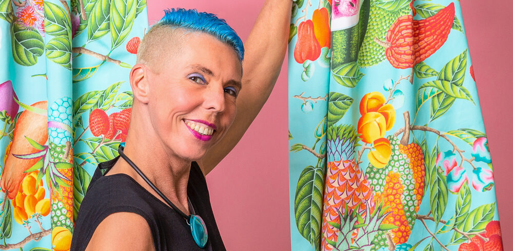 Lachende Person mit kurzen, leuchtend blauen Haaren und einem pinken Lippenstift, die in einem schwarzen Oberteil hinter zwei Vorhängen mit einem auffälligen, bunten, tropischen Fruchtmuster steht und diese zur Seite zieht.