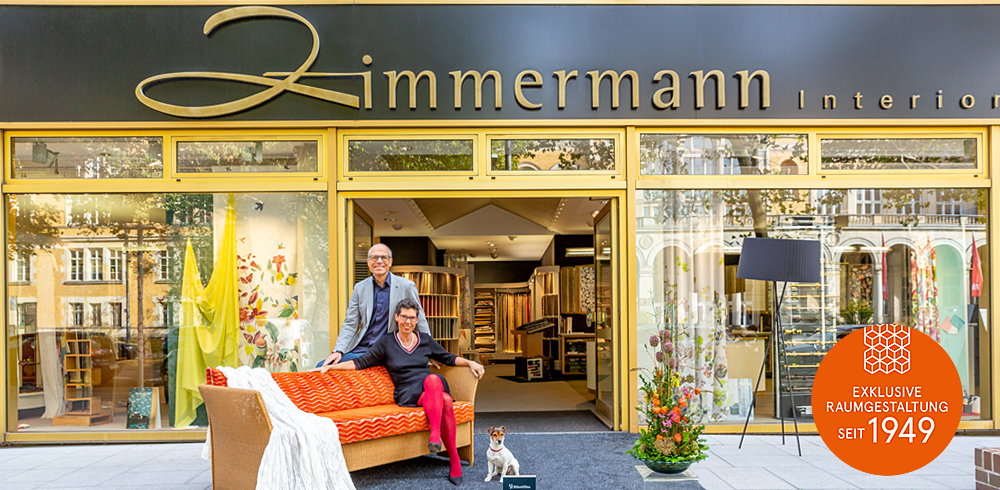 ladenfront Zum Jubiläum Zimmermann Interior Design Straßenansicht des Showrooms. Vor dem Geschäft auf einem Sofa sitzt die Inhaberin. Dahinter auf der Rückenlehne sitzend der Inhaber. Recht neben der Couch ein kleiner weißer Hund.