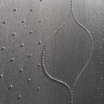 Detailaufnahme einer Tapete von Marburg Colani Evolution. Silbergrauer strukturierter Untergrund wellenförmig verziert mit cremefarbenen Perlen.