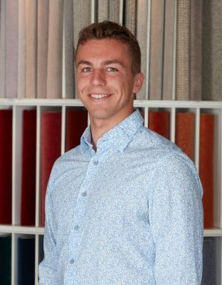 Portrait von Benjamin Zimmermann, Mitarbeiter von Zimmermann InteriorDesign im Bereich EDV-Betreuung. Er steht vor einem bunten teppichmusterregal und trägt ein hellblaues gemustertes Hemd.