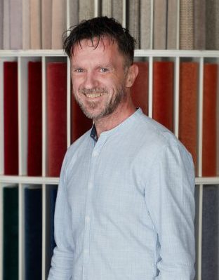 Portrait von Mario Breßler, Mitarbeiter von Zimmermann InterioDesign im Bereich Auftragsabwicklung und technische Produktberatung. Er steht vor einem bunten Teppichmusterregal und trägt ein hellblaues Hemd.