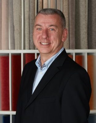 Portrait von Thomas Gericke, Mitarbeiter von Zimmermann InteriorDesign im Bereich Textile Einrichtungsberatung und Umsetzung. Er steht vor einem bunten Teppichmusterregal und trägt ein hellblaues Hemd und schwarzes Jackett.