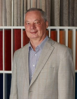 Portrait von Dr. Wolfgang Böhmer, Mitarbeiter von Zimmermann InteriorDesign um Bereich Buchhaltung. Er steht vor einem bunten Teppichmusterregal und trägt ein Hemd mit Jackett darüber.