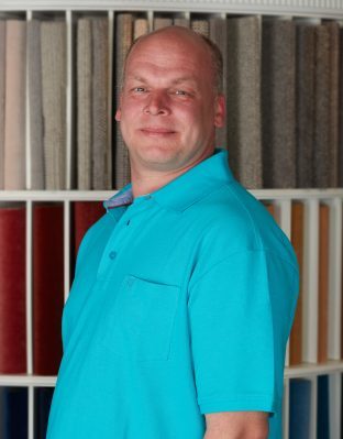 Portrait von Rene Straube, Mitarbeiter von Zimmermann InteriorDesign, zuständig für Silent Gliss Werkstatt und Lieferung. Er trägt ein hellblaues Polo-Shirt und steht vor einem Regal voll Bodenbelagsmustern.