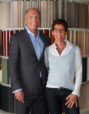 Babette Zimmermann und Steffen Muth-Zimmermann, Geschäftsleitung von Zimmermann InteriorDesign stehen lächelnd im Showroom vor Teppichmustern.